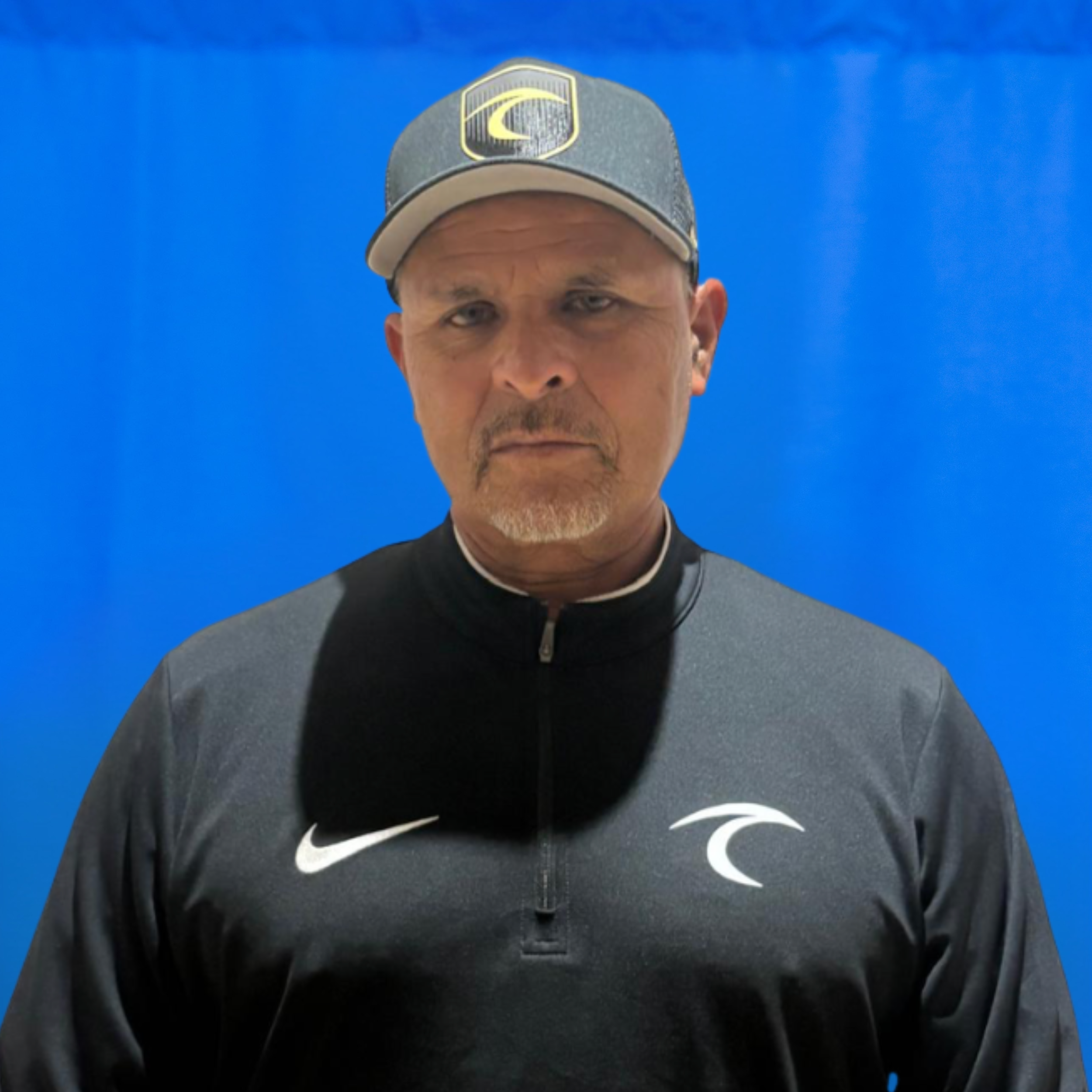 Lorenzo Vazquez Man wearing a dark jacket with a white Nike swoosh and a gray cap with a yellow emblem, against a blue background, portrait-style.