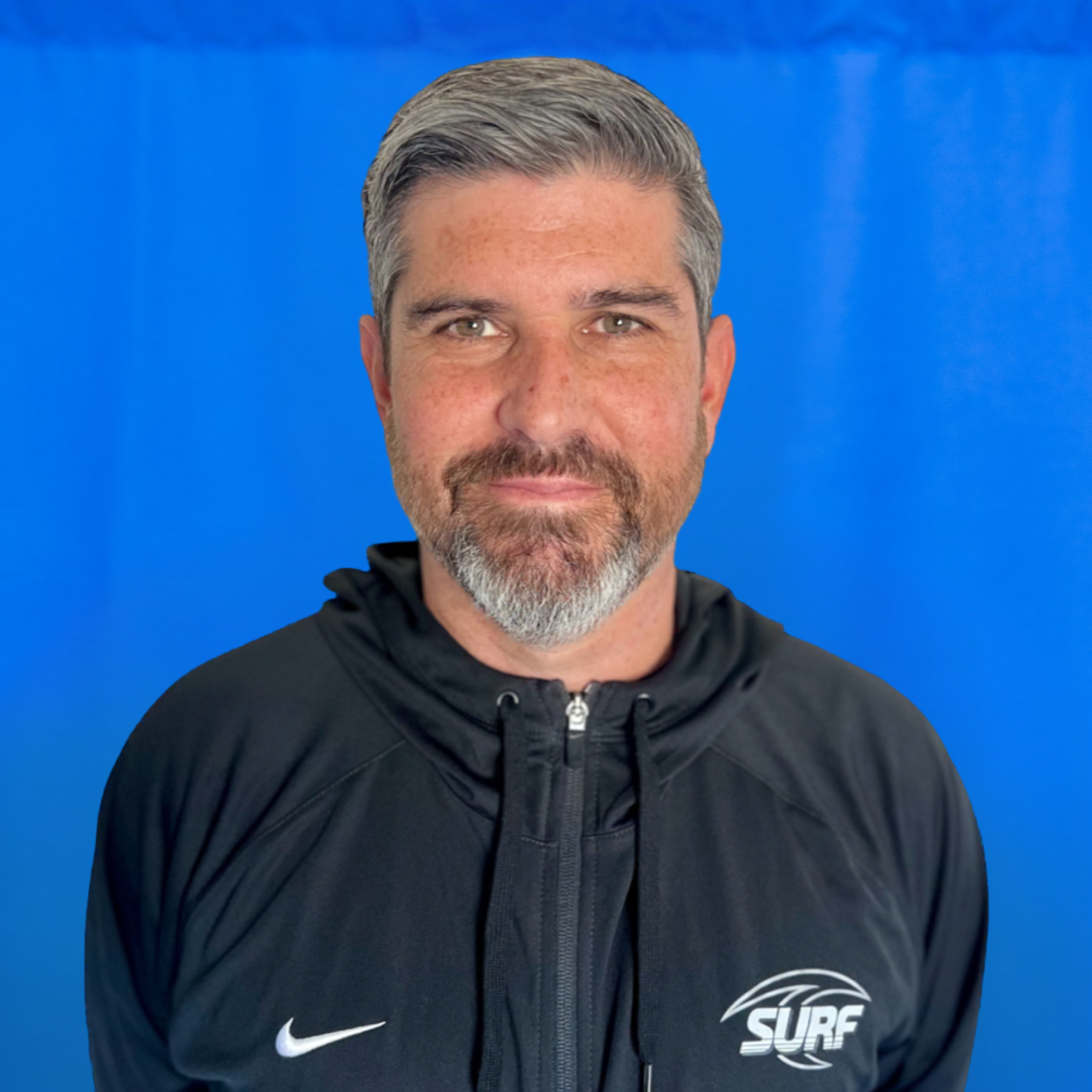 Shawn Ochoa Close-up portrait of a person wearing a black zip-up jacket with a white Nike swoosh on the left chest and a Surf logo on the right against a bright blue background